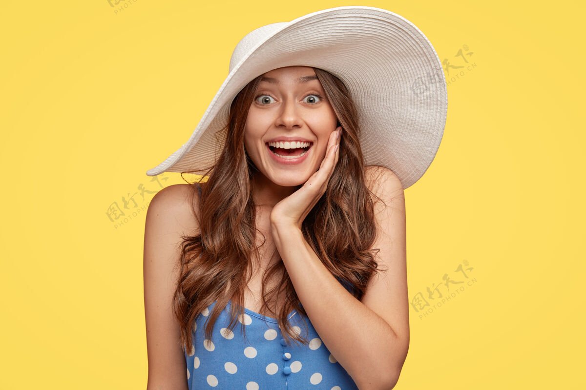 年轻惊艳微笑的年轻女士有着自然的黑发 洁白的牙齿 灿烂的笑容 用手抚摸脸颊 戴着时髦的夏日帽子 对伟大的新闻感到惊讶 隔着黄色的墙女性化惊人帽子