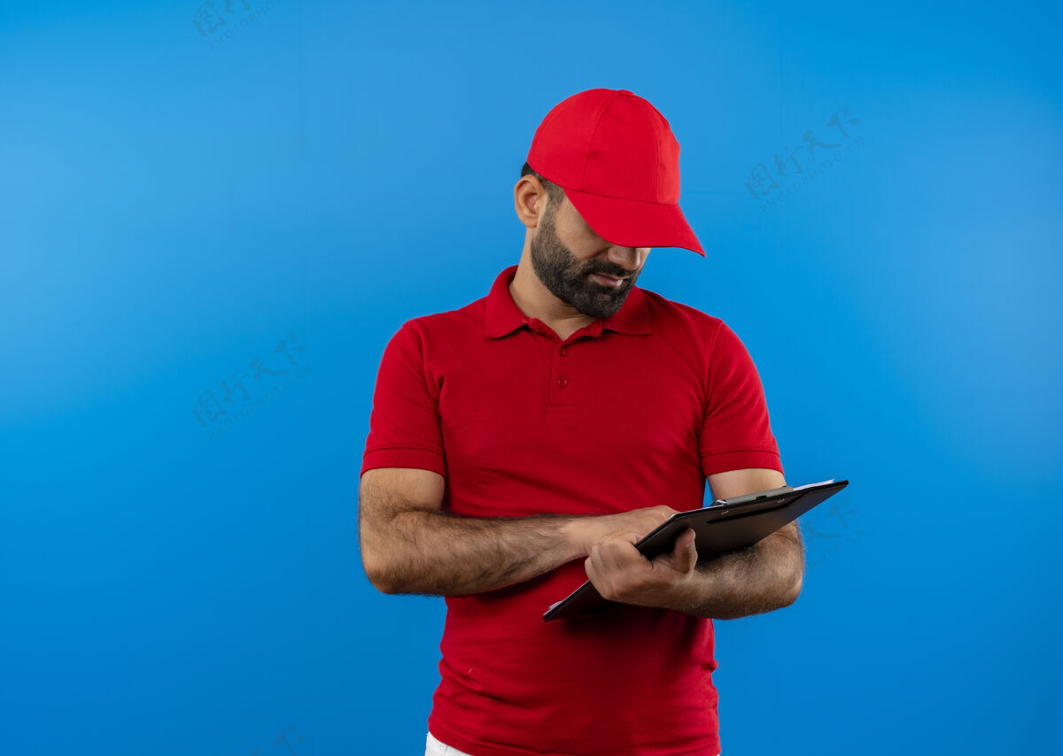 严肃留着胡子的送货员穿着红色制服 戴着帽子 手里拿着剪贴板 手里拿着空白页 严肃地站在蓝色的墙上看着他们递送男人空白