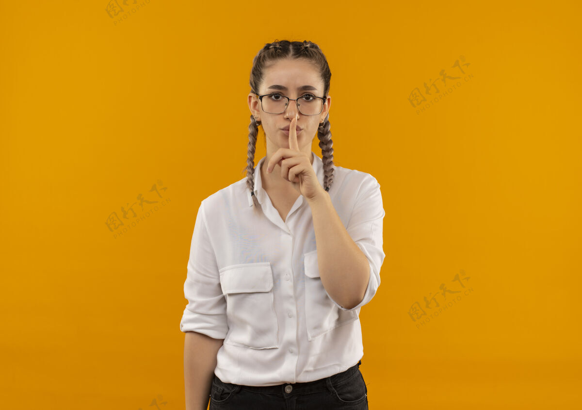 年轻人戴着眼镜 梳着辫子 穿着白衬衫的年轻女学生站在橙色的墙上 朝前面做着沉默的手势看嘴唇公民