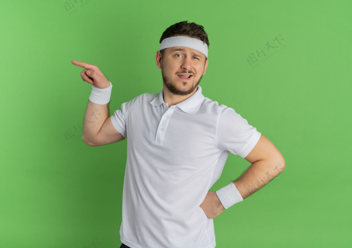 公民身穿白衬衫 头箍朝前看的年轻健身男子 手指指向站在绿色墙壁上的一侧手势姿势头带