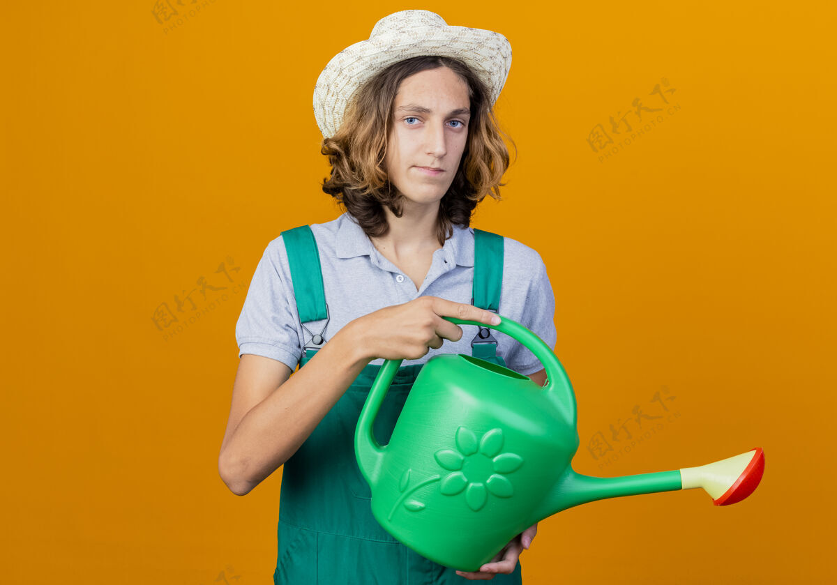 微笑年轻的园丁穿着连体衣戴着帽子拿着浇水罐抱着男人连身衣