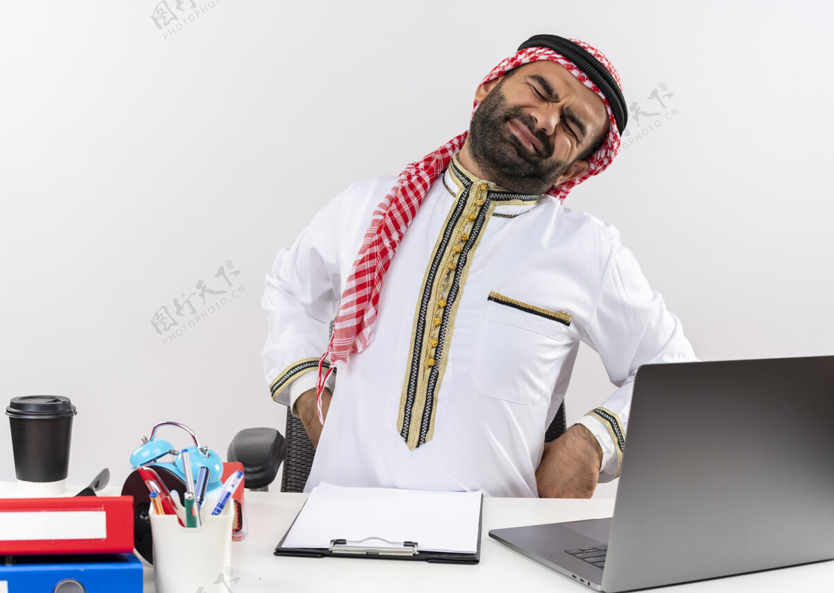 笔记本电脑身着传统服装的阿拉伯商人坐在桌旁 手提电脑看起来很累 在办公室工作时摸背很痛电脑传统坐