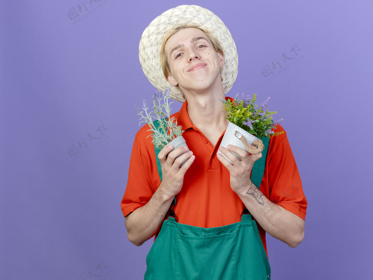 脸年轻的园丁 穿着连体衣 戴着帽子 手里拿着盆栽植物站相机植物