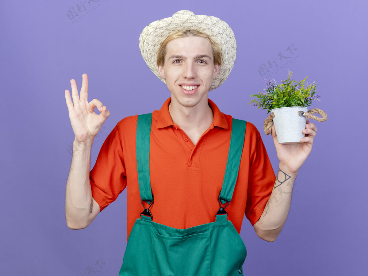 抱着年轻的园丁穿着连体衣 戴着帽子 手里拿着盆栽植物微笑着相机标志站着