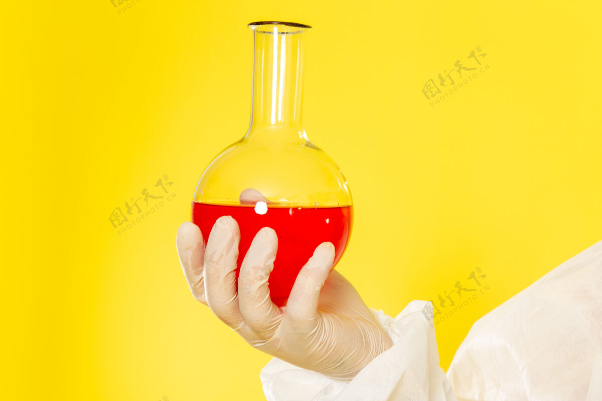 西装正面近距离观察穿着特殊防护服的男性科学工作者在黄色桌子上拿着装有红色溶液的烧瓶化学伏特加特殊
