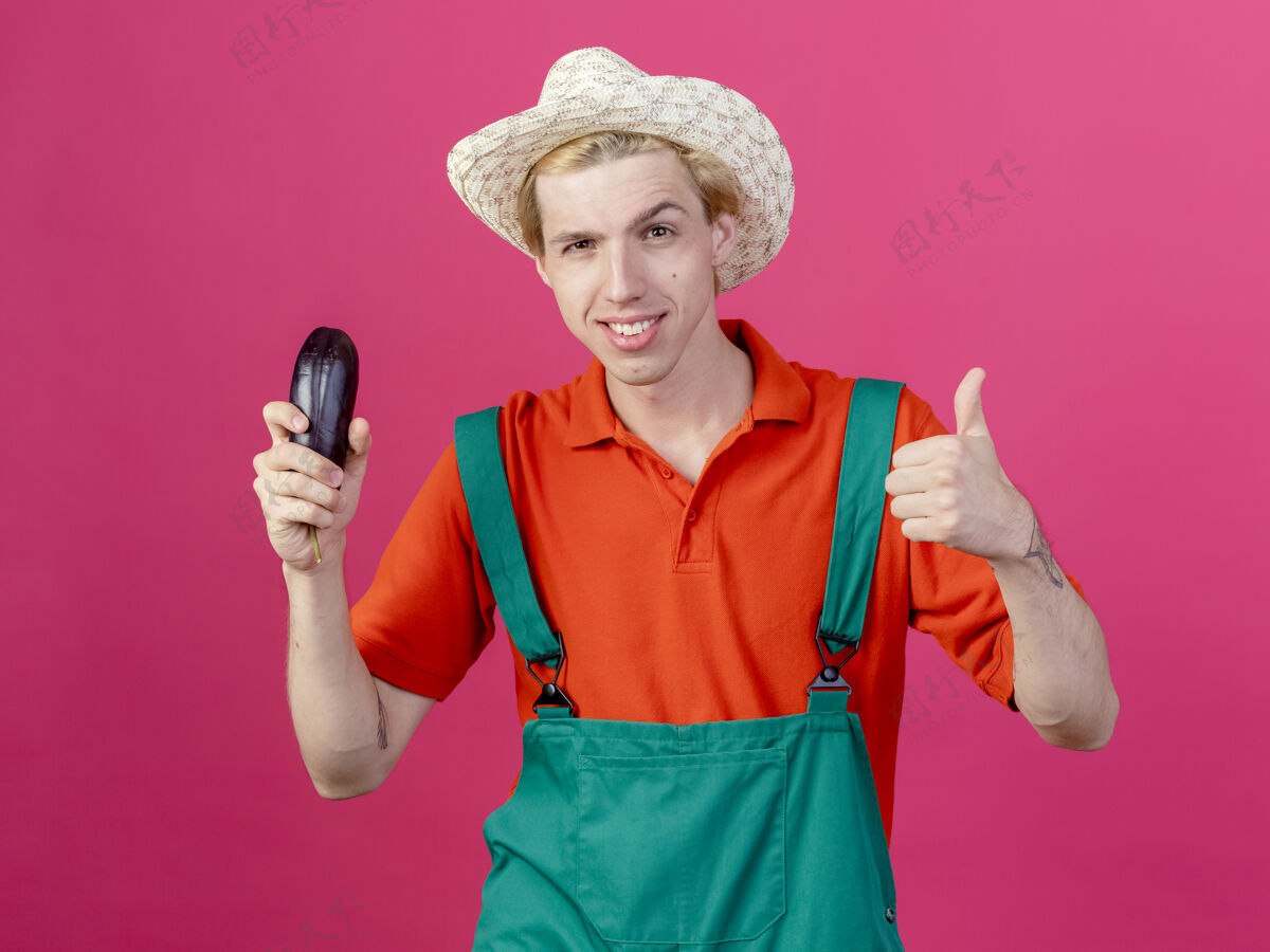 穿年轻的园丁穿着连体衣戴着帽子拿着茄子微笑着竖起大拇指男人拇指年轻人