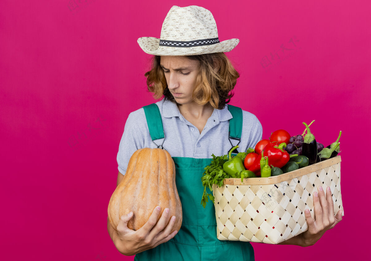 男人年轻的园丁穿着连体衣戴着帽子拿着装满新鲜蔬菜的箱子板条箱帽子持有