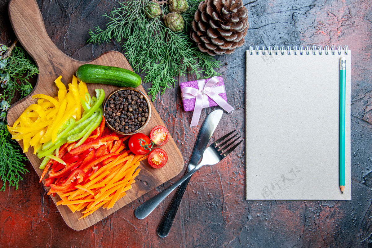 小顶视图彩色切胡椒黑胡椒西红柿黄瓜在砧板上铅笔在记事本上刀叉小礼物在深红色的桌子上番茄顶部蔬菜