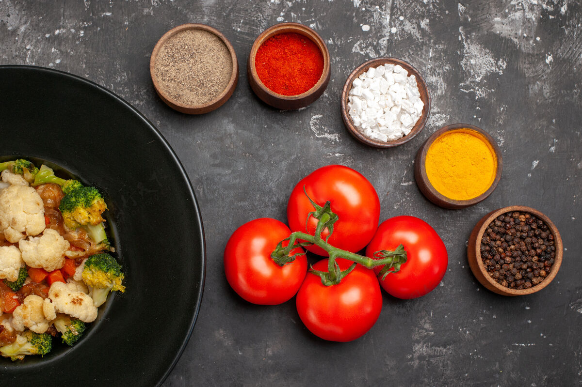 盘子俯视图西兰花和花椰菜沙拉在黑色椭圆形盘子上不同的香料西红柿在黑暗的表面顶部樱桃番茄李子番茄