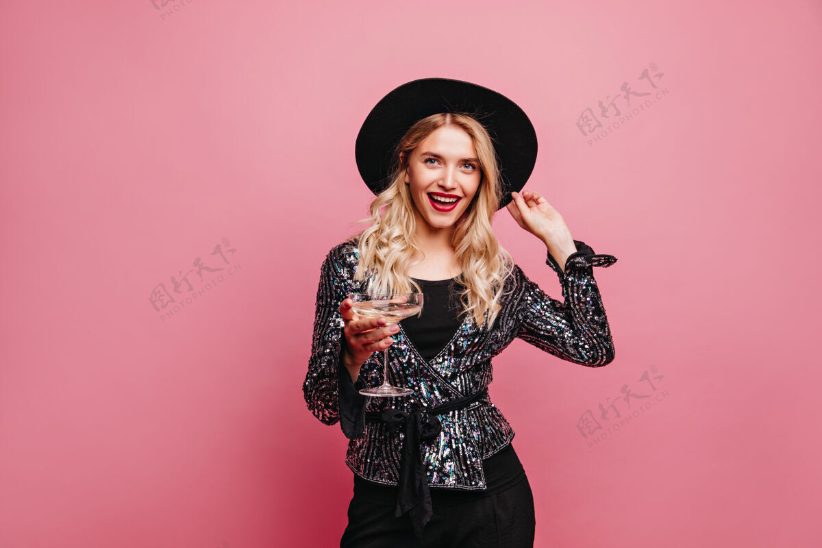 寒冷迷人的白人妇女与酒杯在粉彩墙上摆姿势幽默的金发女孩戴着黑色帽子的室内照片情绪表情放松