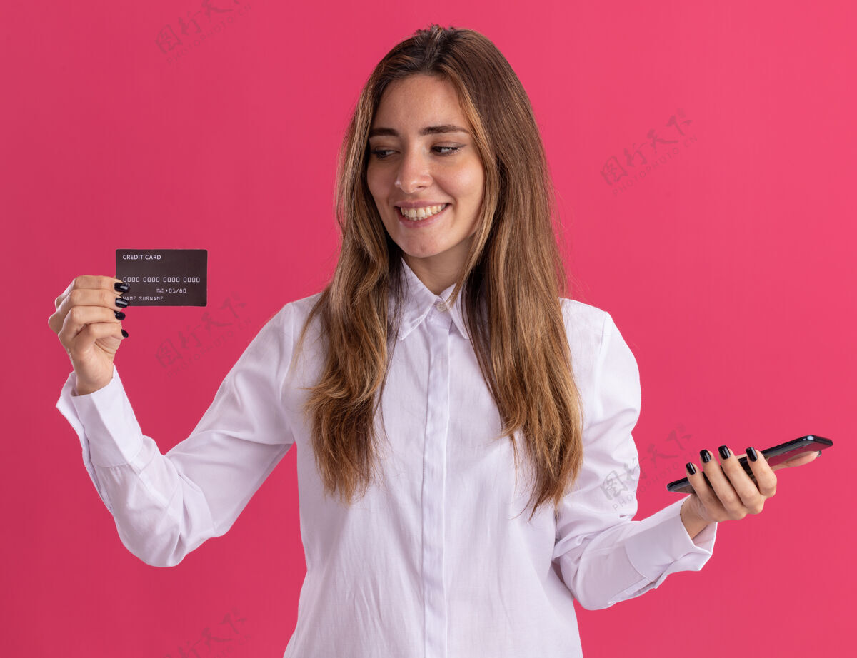 信用微笑着的年轻漂亮的白人女孩拿着电话 看着隔离在粉色墙上的信用卡 还有复印空间漂亮电话年轻