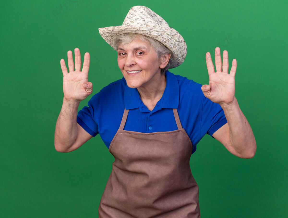 微笑微笑的上了年纪的女园丁戴着园艺帽 四个手指孤立地在绿色的墙上做着手势 还有复制空间戴手势园艺