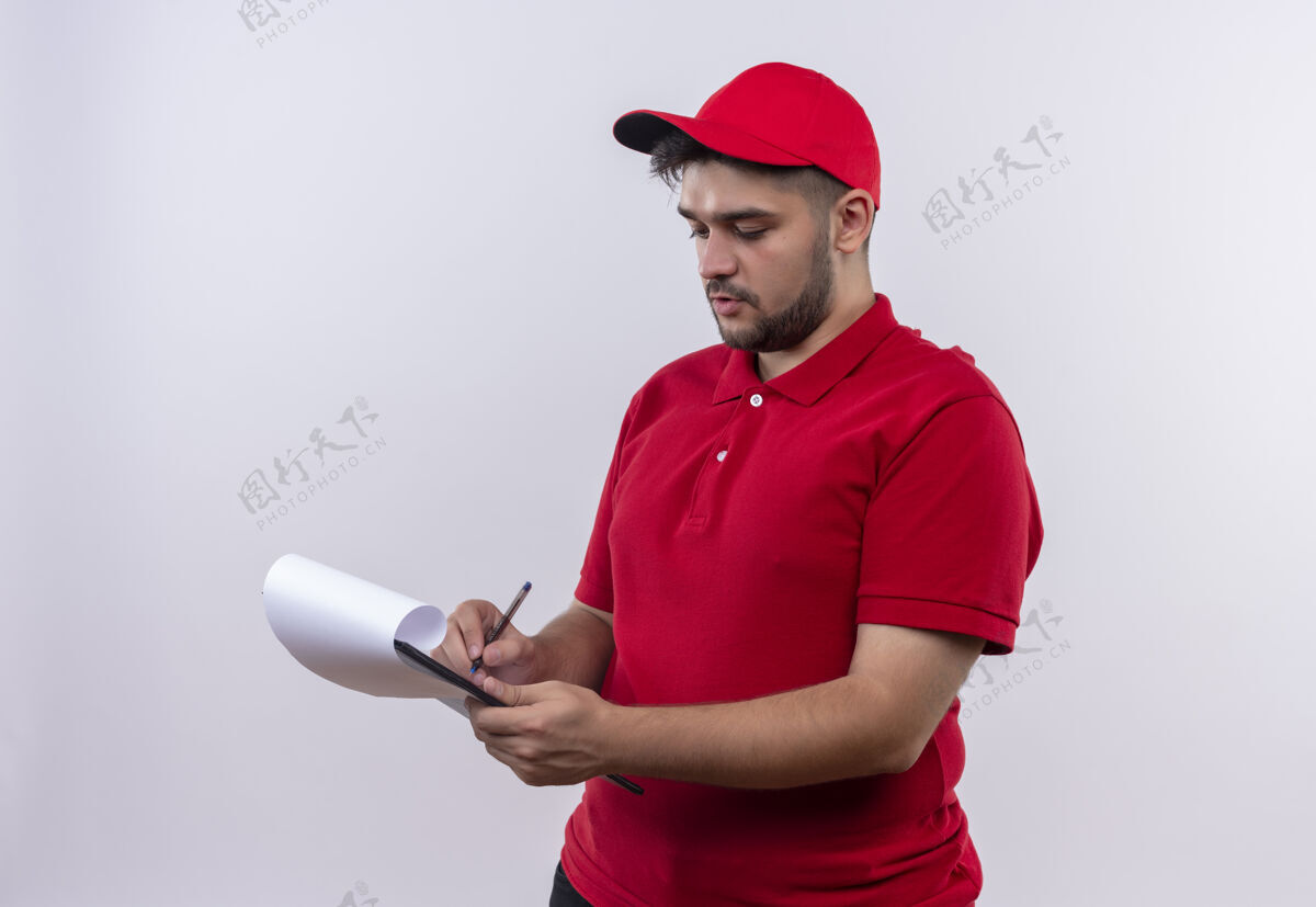 站年轻的送货员穿着红色制服 戴着帽子 严肃地看着写字板上的写字板看木板男
