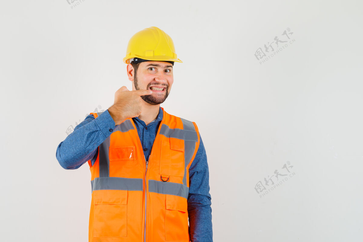 勤杂工年轻的建筑工人指着他的牙齿在衬衫 背心 头盔和看起来愉快 前视图年轻人工具工厂