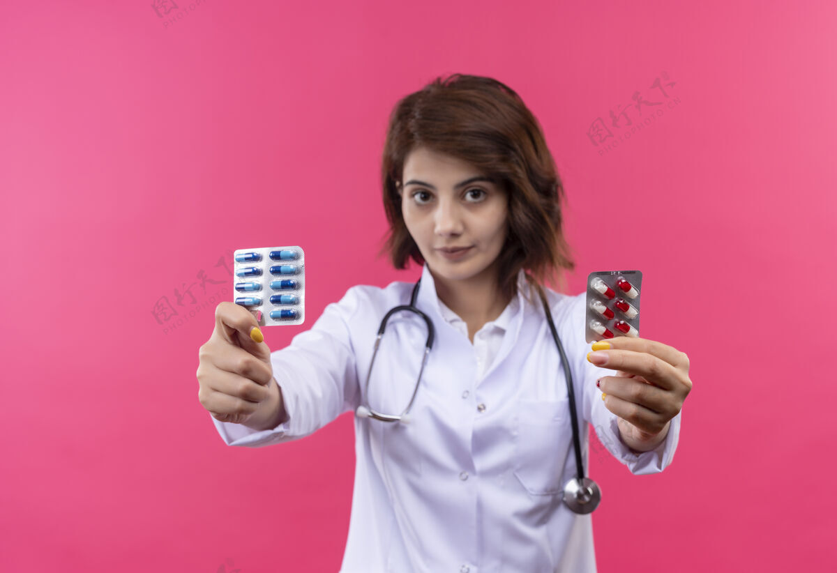 支架身穿白大褂 手持听诊器的年轻女医生站在粉红色的墙上 露出水泡和药丸女人水泡年轻人