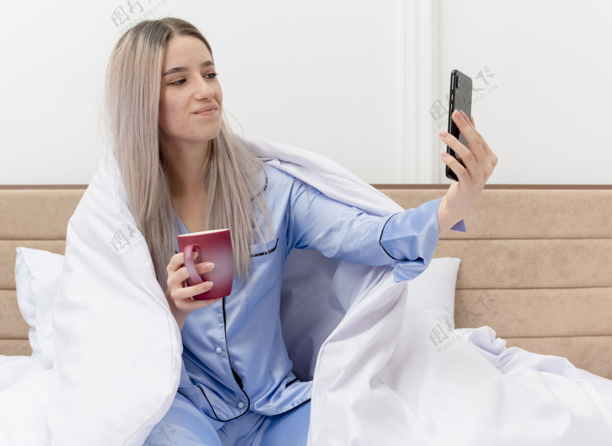 咖啡穿着蓝色睡衣的年轻美女坐在床上 喝着咖啡 在明亮的背景下 用智能手机在卧室里快乐而积极地自拍室内床睡衣