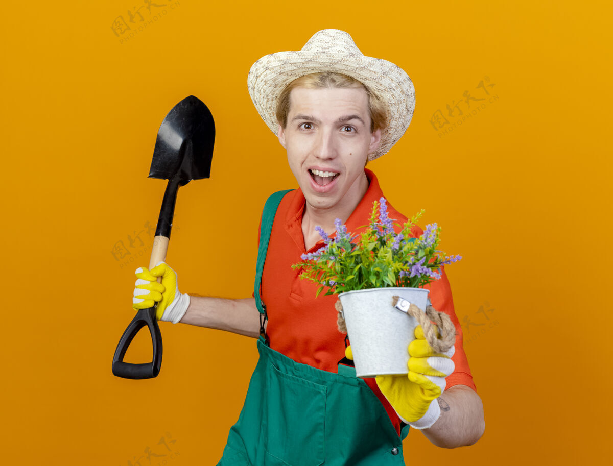 帽子年轻的园丁穿着连体衣 戴着帽子 拿着铲子 展示着盆栽植物 站在橙色的背景上开心地微笑着看着镜头锅铲子欢呼