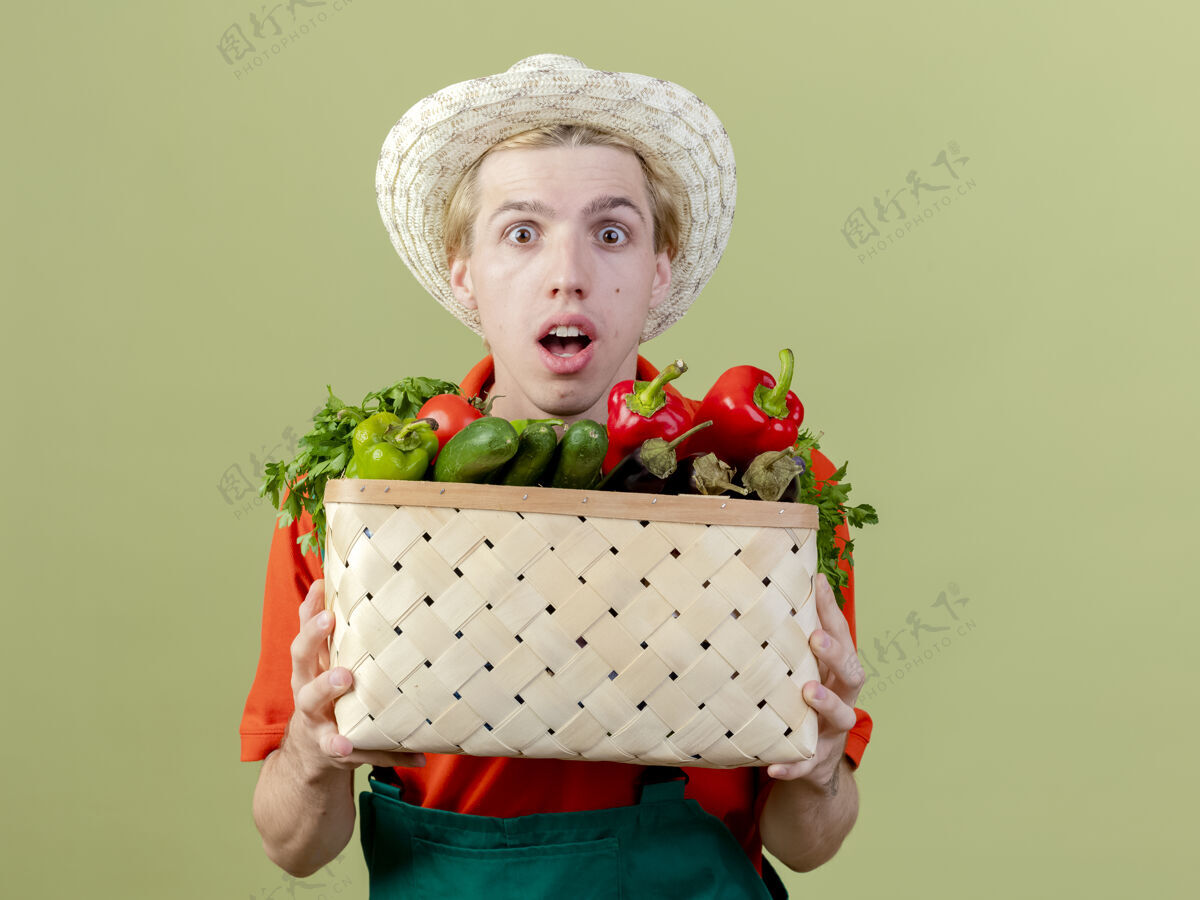 板条箱年轻的园丁穿着连体衣 戴着帽子 手里拿着装满蔬菜的箱子 站在灯光的背景下看着摄像机 既惊讶又惊讶男人看惊喜