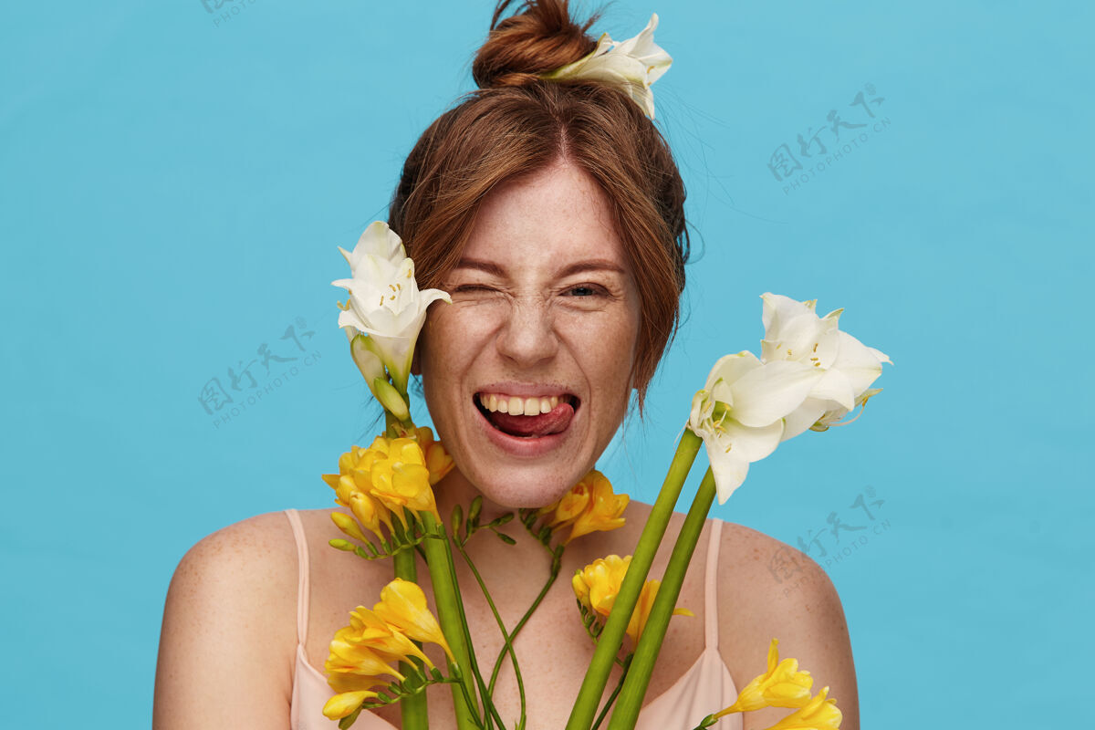 舌头有趣的镜头年轻迷人的红发女士与发髻发型显示愉快的舌头 而愚弄 站在蓝色背景与春天的花朵微笑发髻肖像