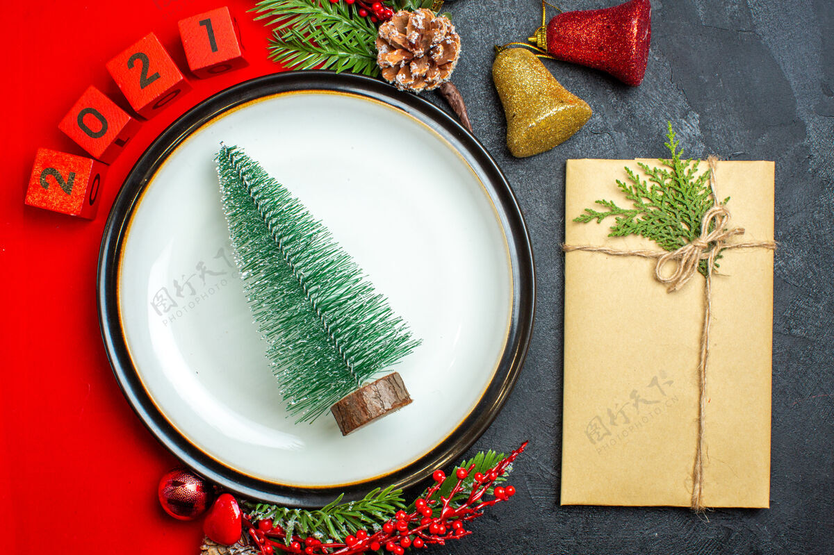 容器新年背景俯视图 圣诞树放在餐盘上装饰配件杉木树枝和数字放在红色餐巾旁边 礼物放在黑色桌子上盘子餐盘风景