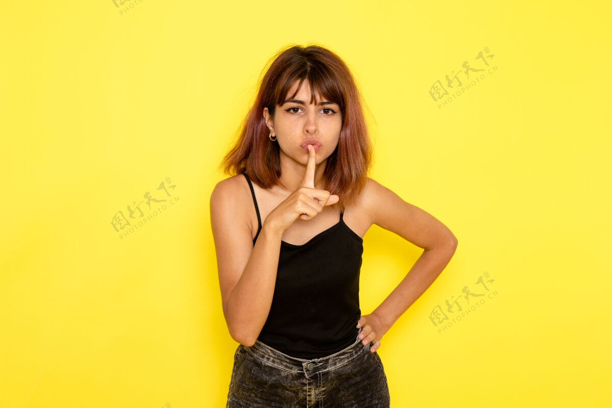 表演身穿黑色衬衫的年轻女性在浅黄色墙壁上摆姿势并显示沉默标志的正面视图女性性感头发