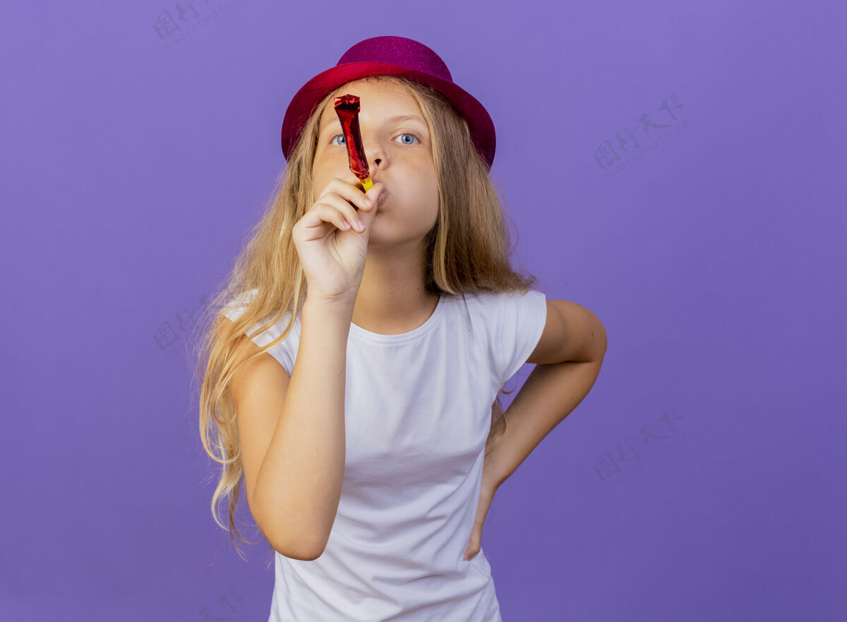 女孩美丽的小女孩在节日帽子吹哨子快乐和积极 生日聚会的概念站在紫色的背景节日派对哨子