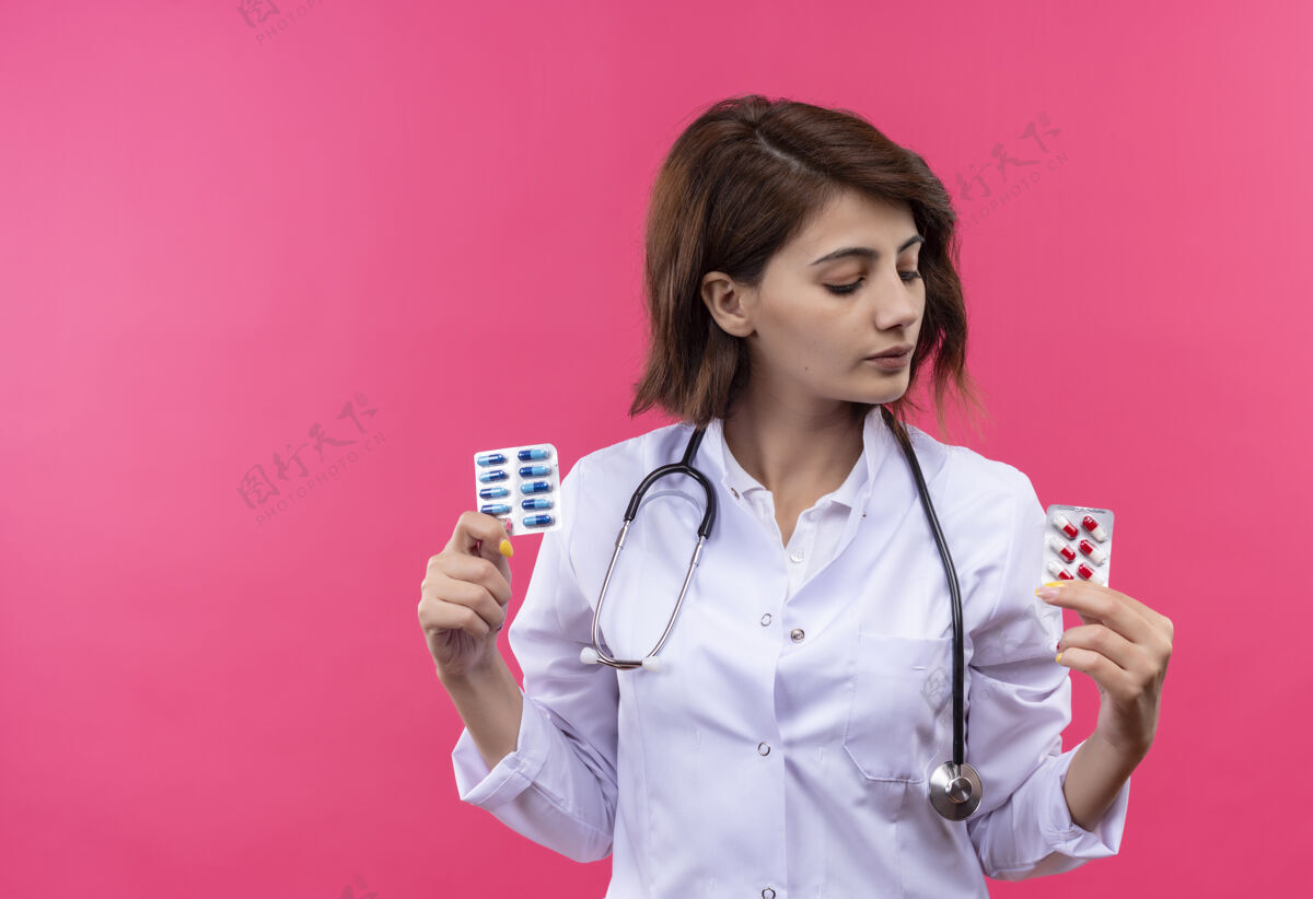 外套年轻的女医生 穿着白大褂 手持听诊器 手里拿着水泡 手里拿着药丸 一脸严肃地看着它粉红严重年轻