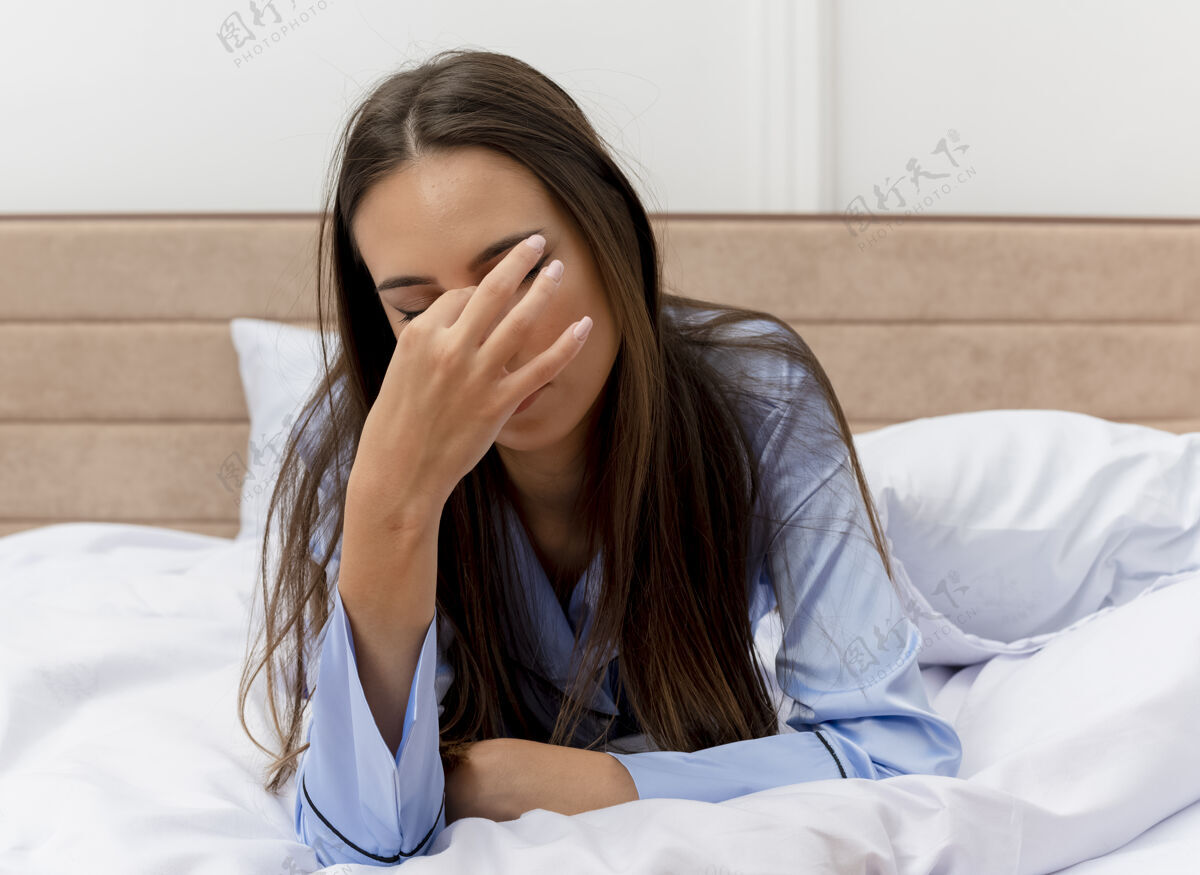 打哈欠穿着蓝色睡衣的年轻美女躺在床上 枕着枕头打呵欠 感觉晨曦中的疲惫在卧室的室内灯光背景下卧室疲劳躺着