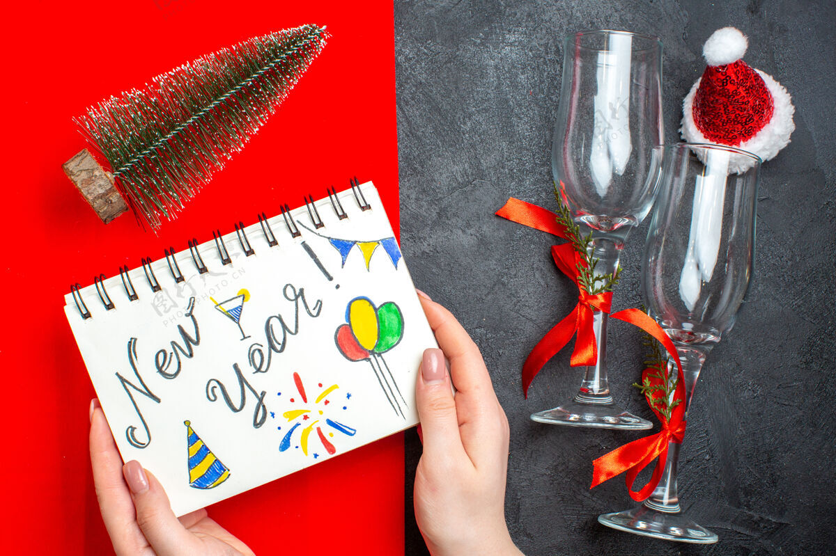 螺旋笔记本手持螺旋笔记本的顶视图 黑色和红色背景上有新年图画和圣诞树玻璃高脚杯手指视图剪刀