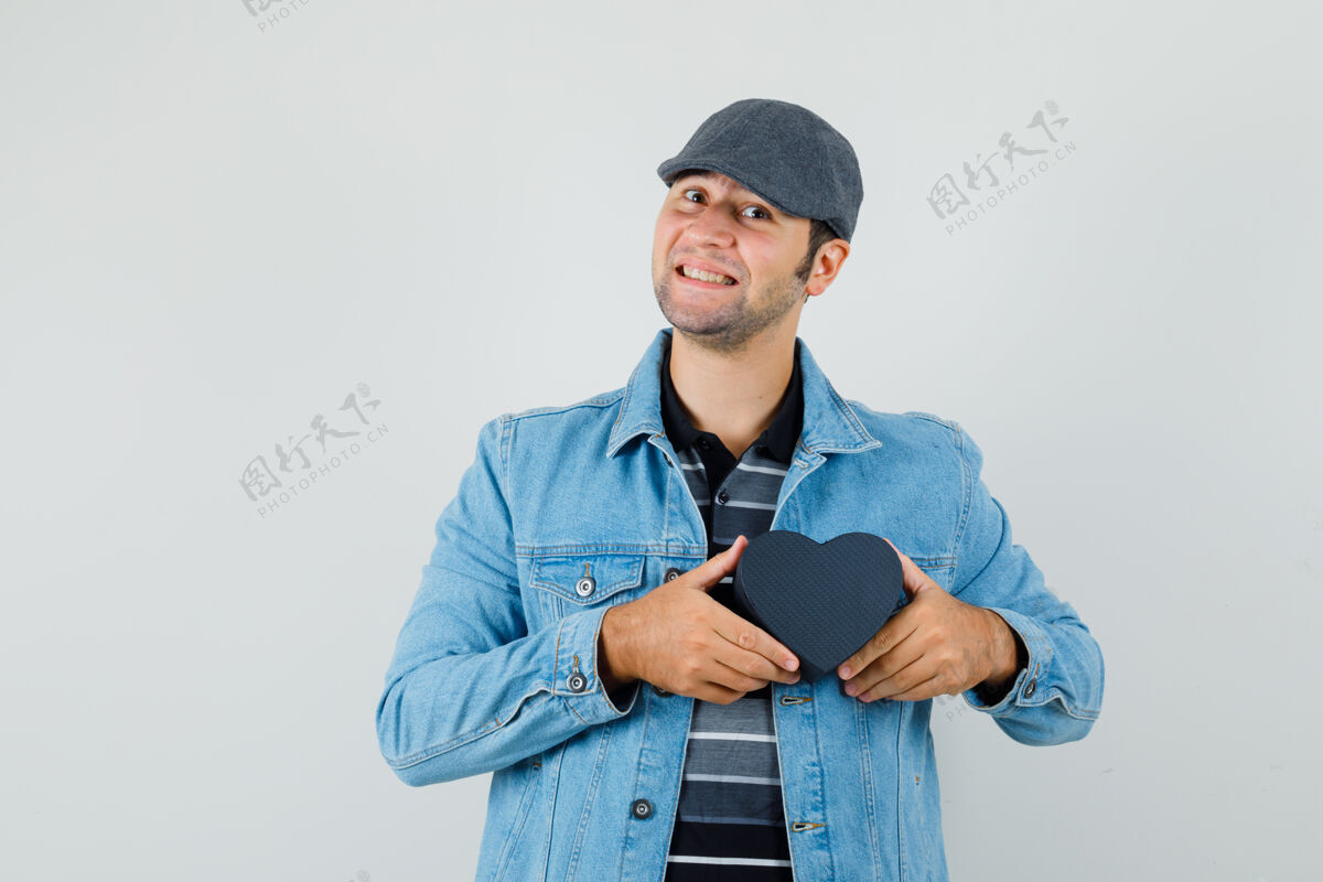 胡茬年轻人穿着夹克 戴着帽子 手里拿着心形盒子 看上去很高兴胡须休闲盒子