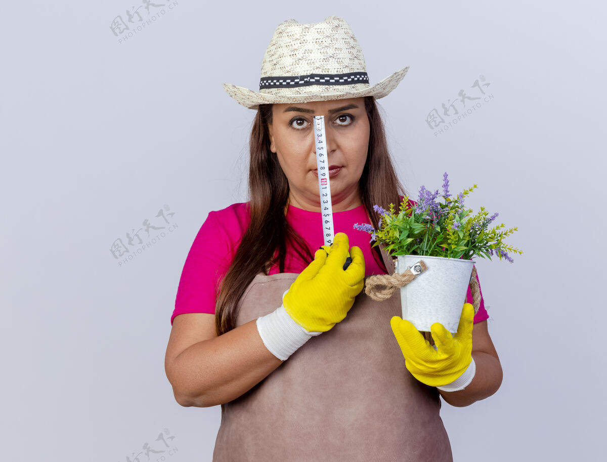 手套穿着围裙 戴着帽子 戴着橡胶手套 拿着盆栽植物和卷尺的中年园丁妇女站在白色背景下严肃地看着摄像机严重穿着举行