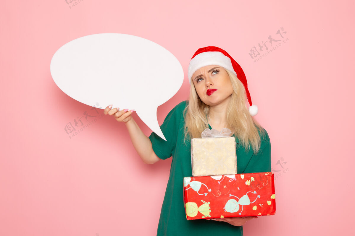 圣诞节正面图年轻女性手持圣诞礼物和白色标志在粉色墙上的女性礼物雪白新年假期微笑帽子美丽