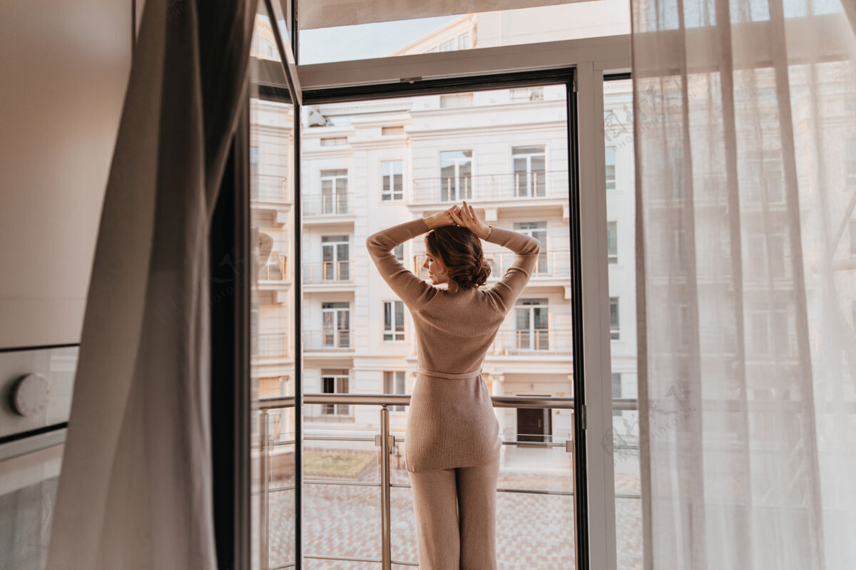 放松一个穿着棕色衣服的放松的女人的画像一个快乐的时尚女士在大窗户边摆姿势的照片放松年轻阳台