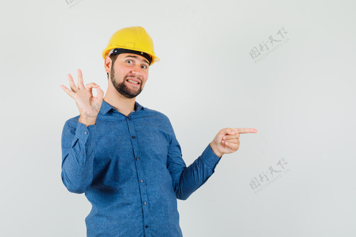 指向年轻的工人指着旁边 在衬衫 头盔上显示ok标志 高兴地看着头盔工业旁听