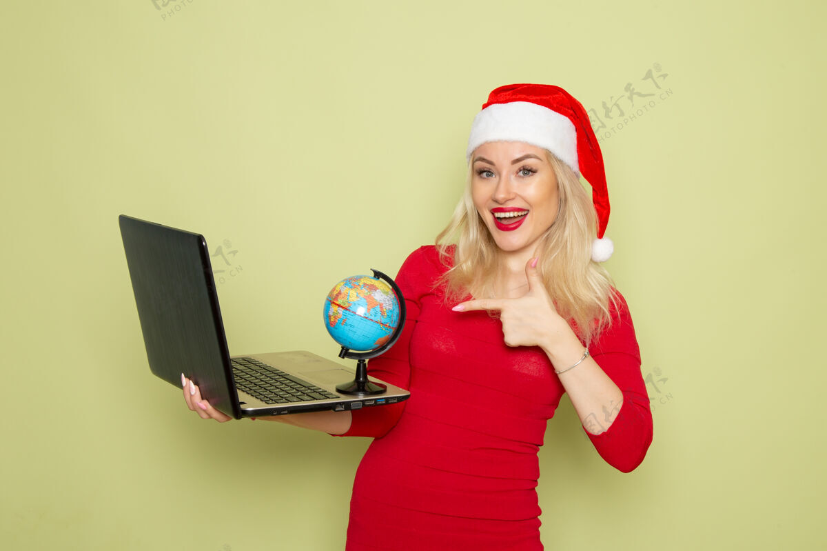 漂亮正面图美丽的女性手持小地球仪 在绿色的墙上用笔记本电脑度假情感圣诞雪新年色彩帽子圣诞节颜色