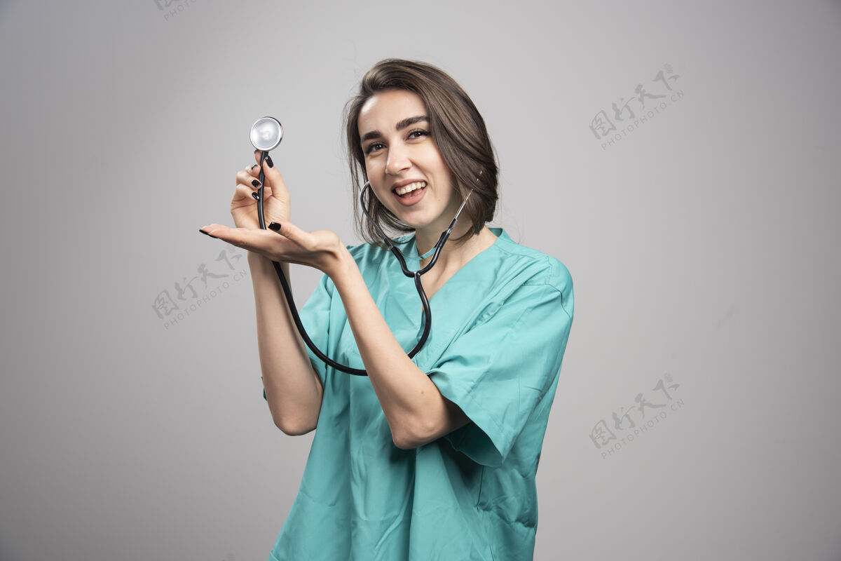 健康快乐的医生在灰墙上展示听诊器职业工作制服