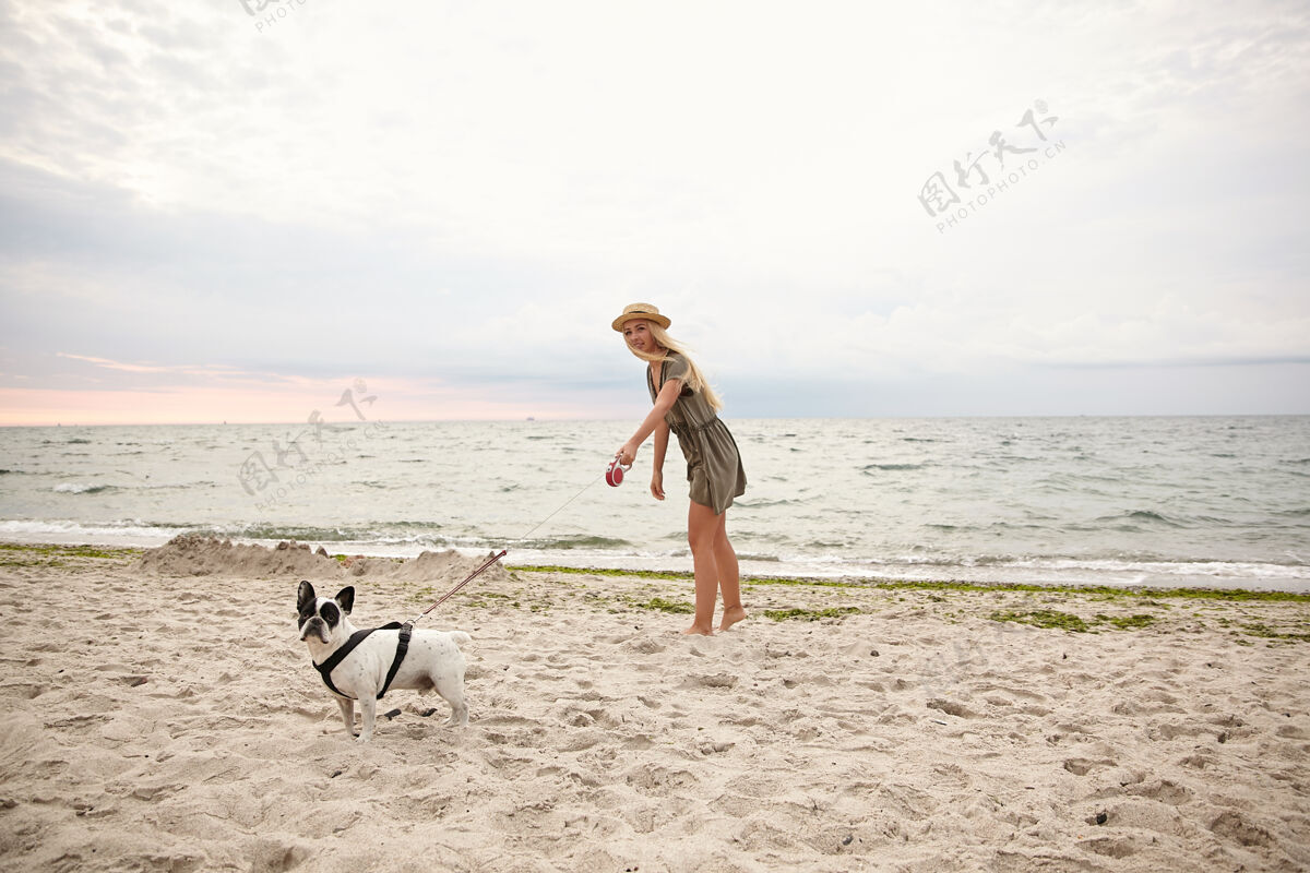 视图一个漂亮的金发年轻女子穿着夏装 戴着草帽 带着她的狗在阴天的沙滩上散步的水平户外镜头多云长白色