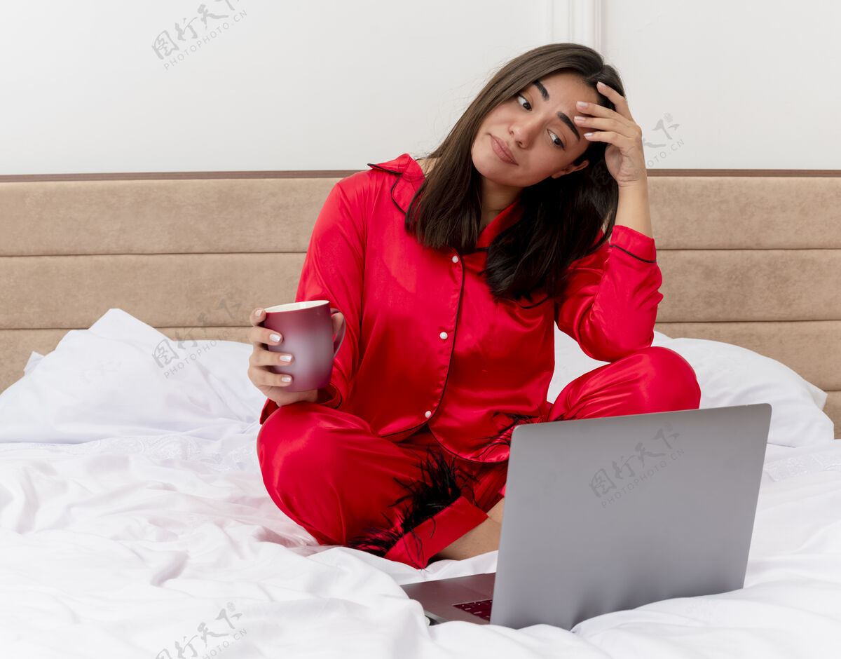 不高兴穿着红色睡衣的年轻漂亮女人坐在床上 手里拿着笔记本电脑和一杯咖啡 在浅色背景下的卧室里显得困惑和不安困惑睡衣咖啡