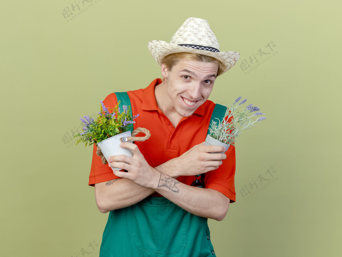 站年轻的园丁穿着连体衣 戴着帽子 手里拿着盆栽植物 微笑着看着镜头 眨着眼睛 站在明亮的背景下 快乐而积极花园男人年轻