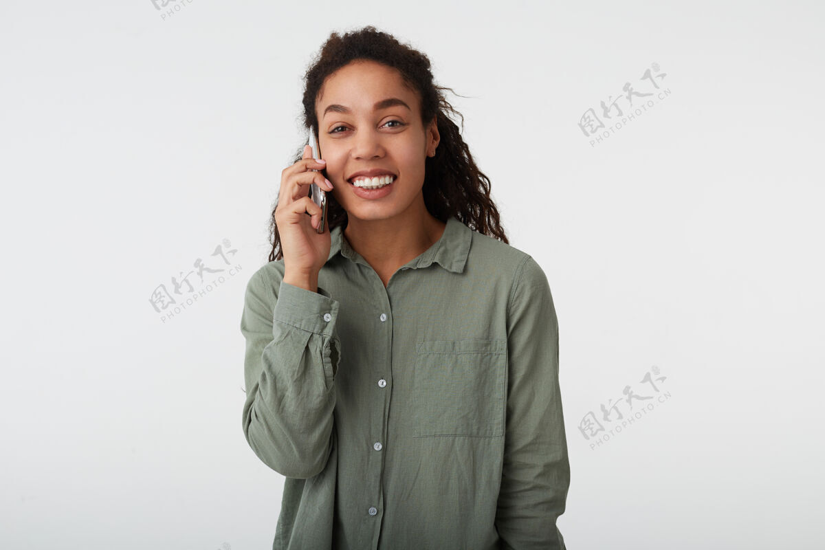 情绪摄影棚拍摄的快乐年轻可爱的黑头发卷曲黑皮肤的女人愉快地微笑着 同时有电话交谈 站在白色背景下的绿色衬衫卷发站立微笑