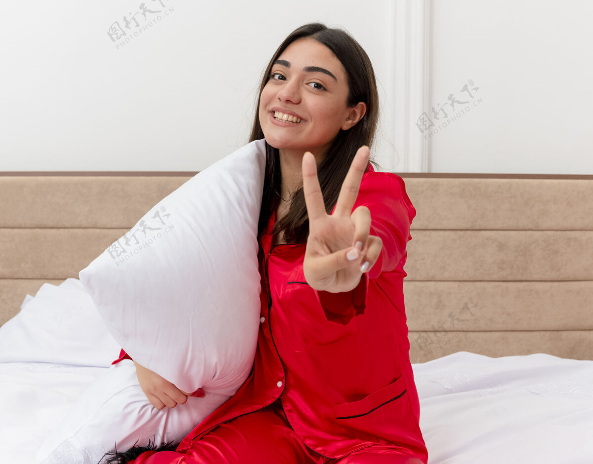 相机穿着红色睡衣的年轻漂亮女人坐在床上 带着枕头看着相机 微笑着在灯光背景下展示卧室内部的v形标志微笑床女人