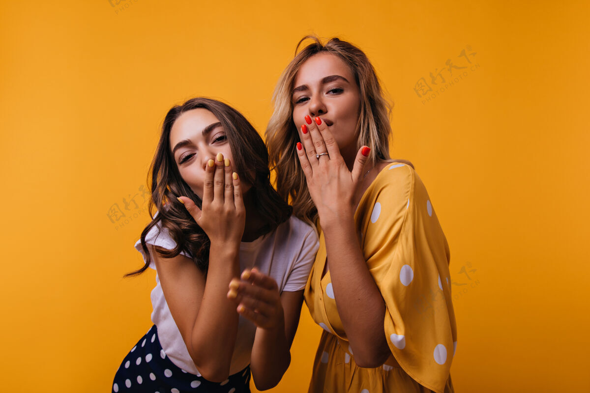 朋友一个棕褐色皮肤的女孩和朋友在黄色的床上摆姿势时送上了飞吻可爱的欧洲姐妹们在一起玩得很开心积极服装女性
