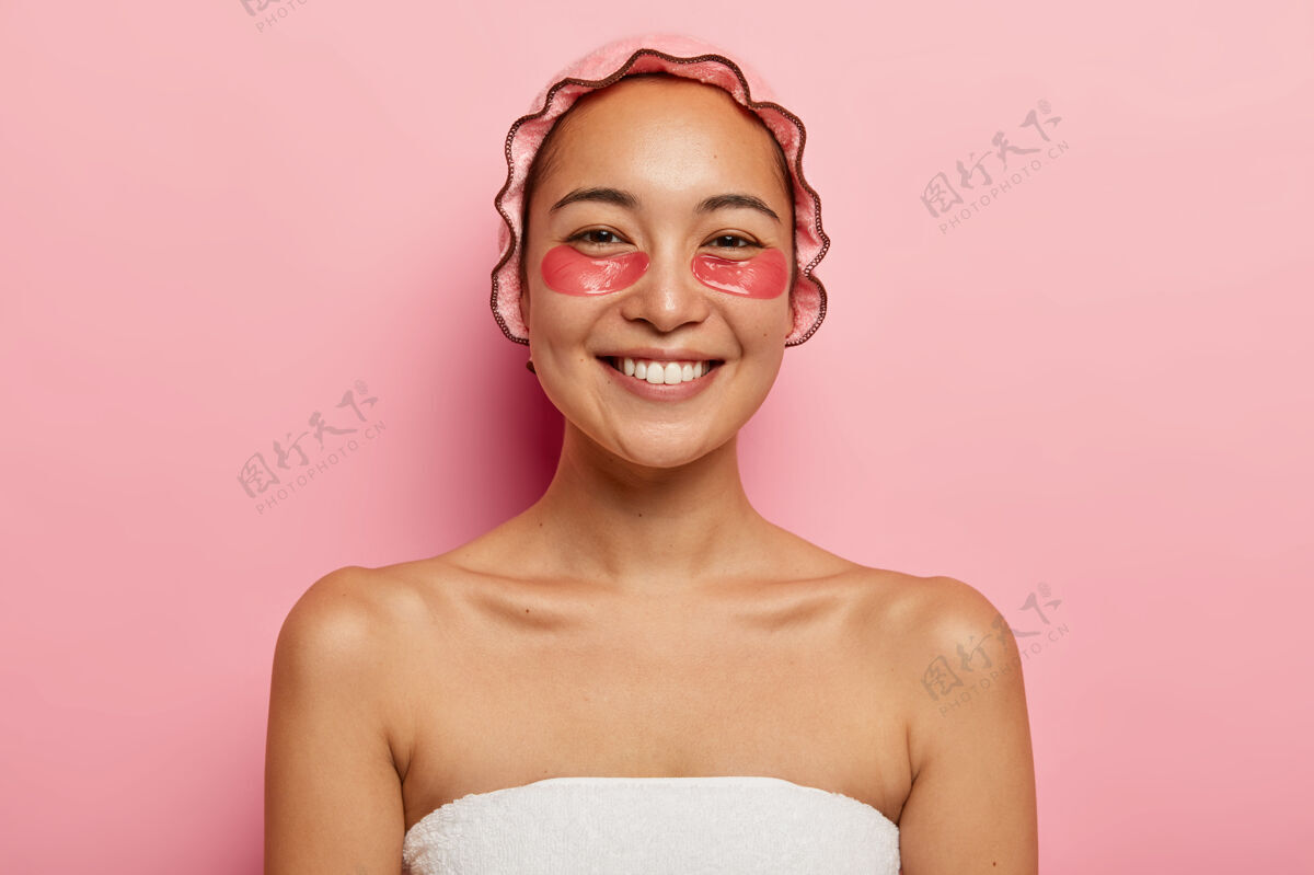 特写美丽微笑的女人的特写肖像有美容治疗 戴粉红色的淋浴帽 裹在毛巾里 眼睛下面有胶原蛋白垫以减少皱纹 站在室内美丽的概念无袖女士垫