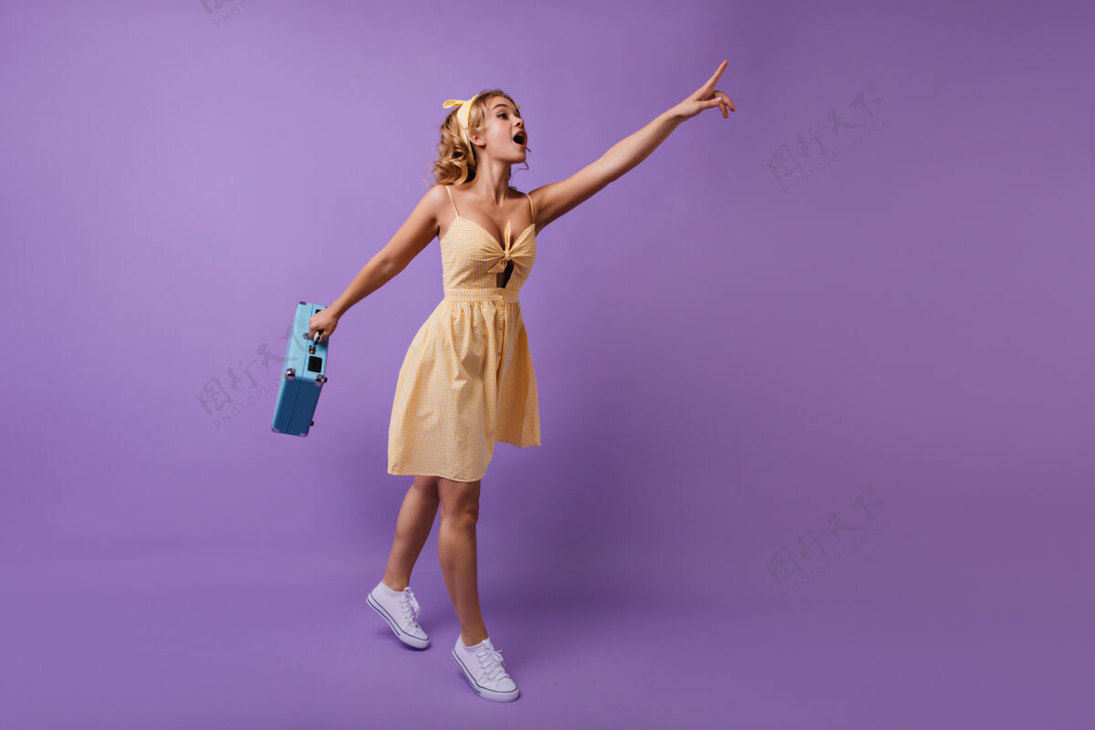 女性热情的女人 手里拿着蓝色的手提箱 手指着什么东西穿着黄色裙子的有趣好奇的女孩的全幅肖像行李包紫色