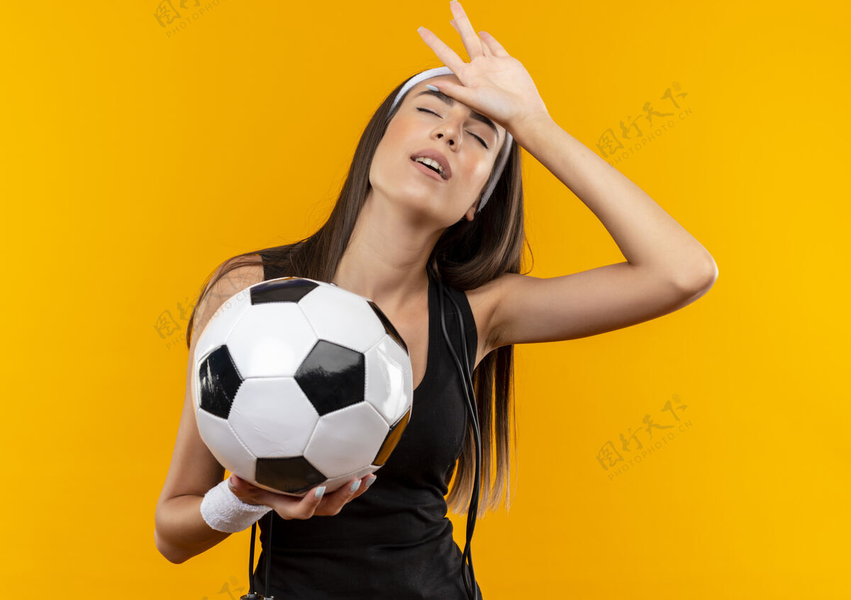 足球疲惫的年轻漂亮的运动女孩戴着头带和腕带拿着足球把手放在头上闭着眼睛跳绳围着脖子孤立在橙色的空间闭上头运动