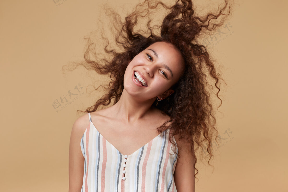 长一幅室内肖像画 欢快的年轻女子挥舞着她卷曲的棕色头发 面带笑容 愉快地看着 摆着米色的姿势黑发女性发型
