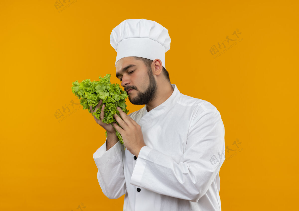 拿着身着厨师制服的年轻男厨师手持和嗅着隔离在橙色空间的莴苣制服嗅着厨师