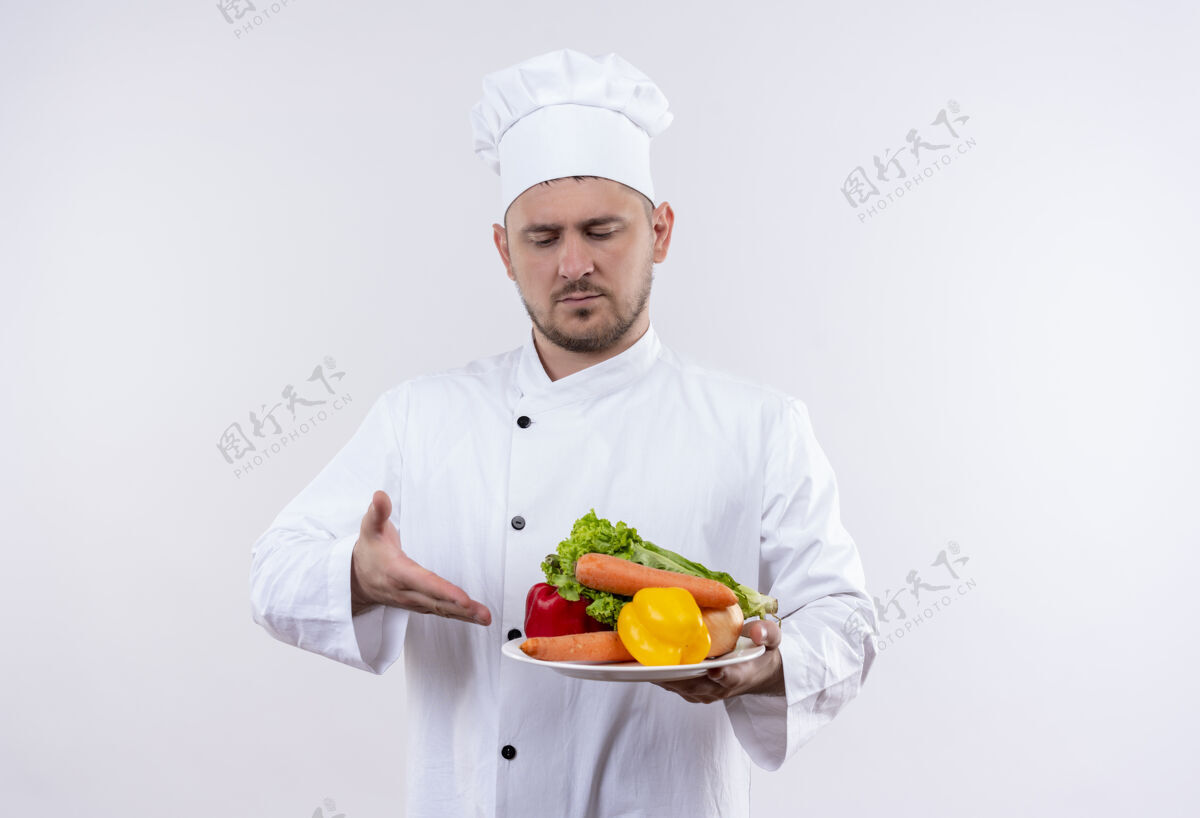 厨师年轻帅气的厨师穿着厨师制服 拿着盘子里的蔬菜 看着并指着孤立的白色空间里的蔬菜年轻盘子看着