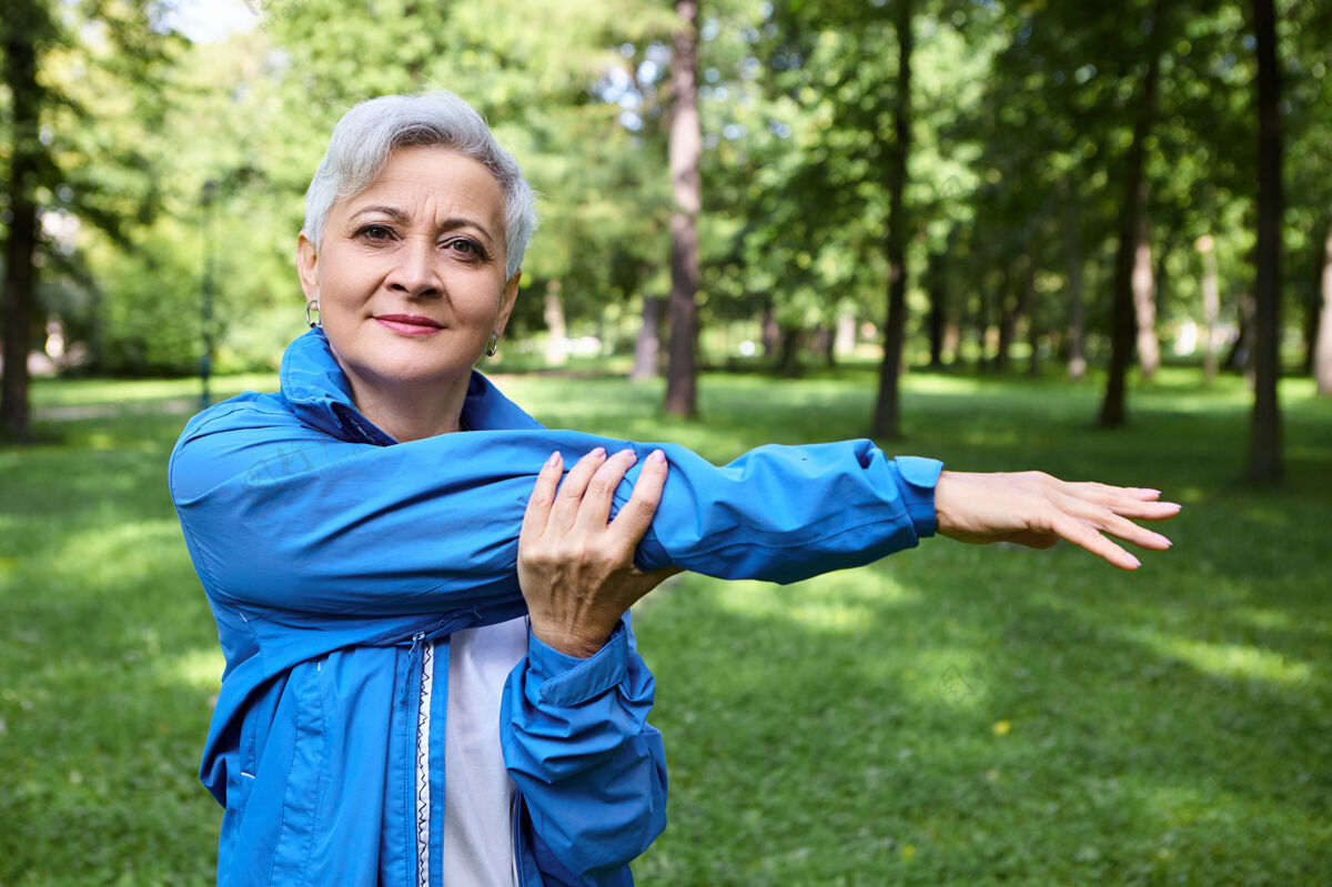 老年人户外拍摄健康运动的老年妇女与灰色短发在公园锻炼老年妇女在蓝色运动夹克伸展手臂肌肉 热身前运行锻炼运动肖像退休