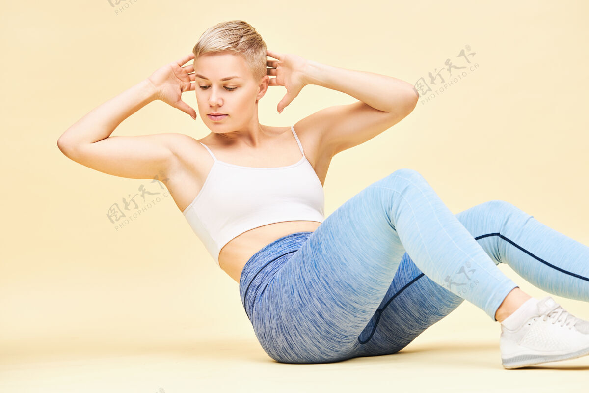 仰卧起坐美丽年轻的欧洲女健身教练 一头小精灵般的金发 双手放在脑后 展示腹肌紧缩或交叉运动的正确技巧 让腹肌得到锻炼运动装腹部健身